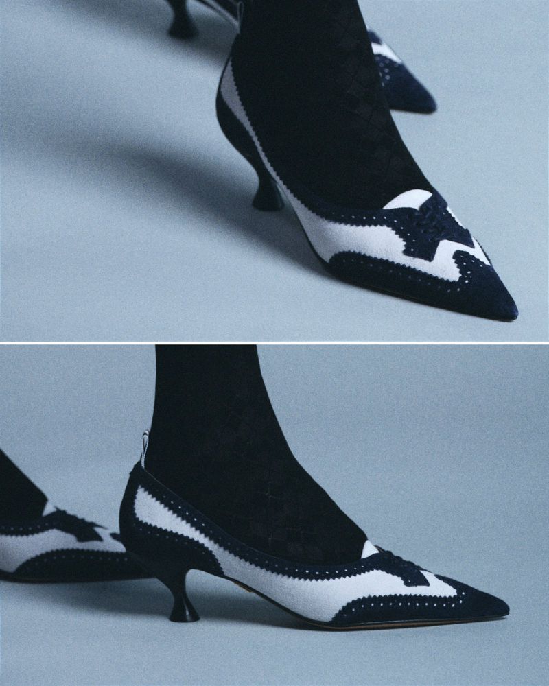 Close-up of two-tone black and white heeled shoes with pointed toes and black hosiery against a light gray backdrop; footwear fashion.