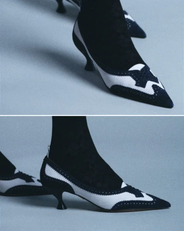 Close-up of two-tone black and white heeled shoes with pointed toes and black hosiery against a light gray backdrop; footwear fashion.