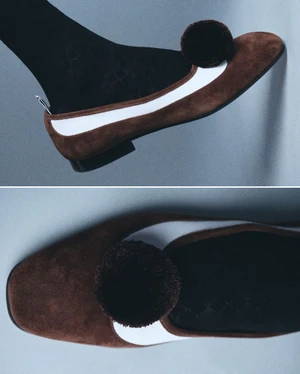 Close-up of brown suede flat shoe with white stripe and pom-pom, worn with black patterned socks against a pale background