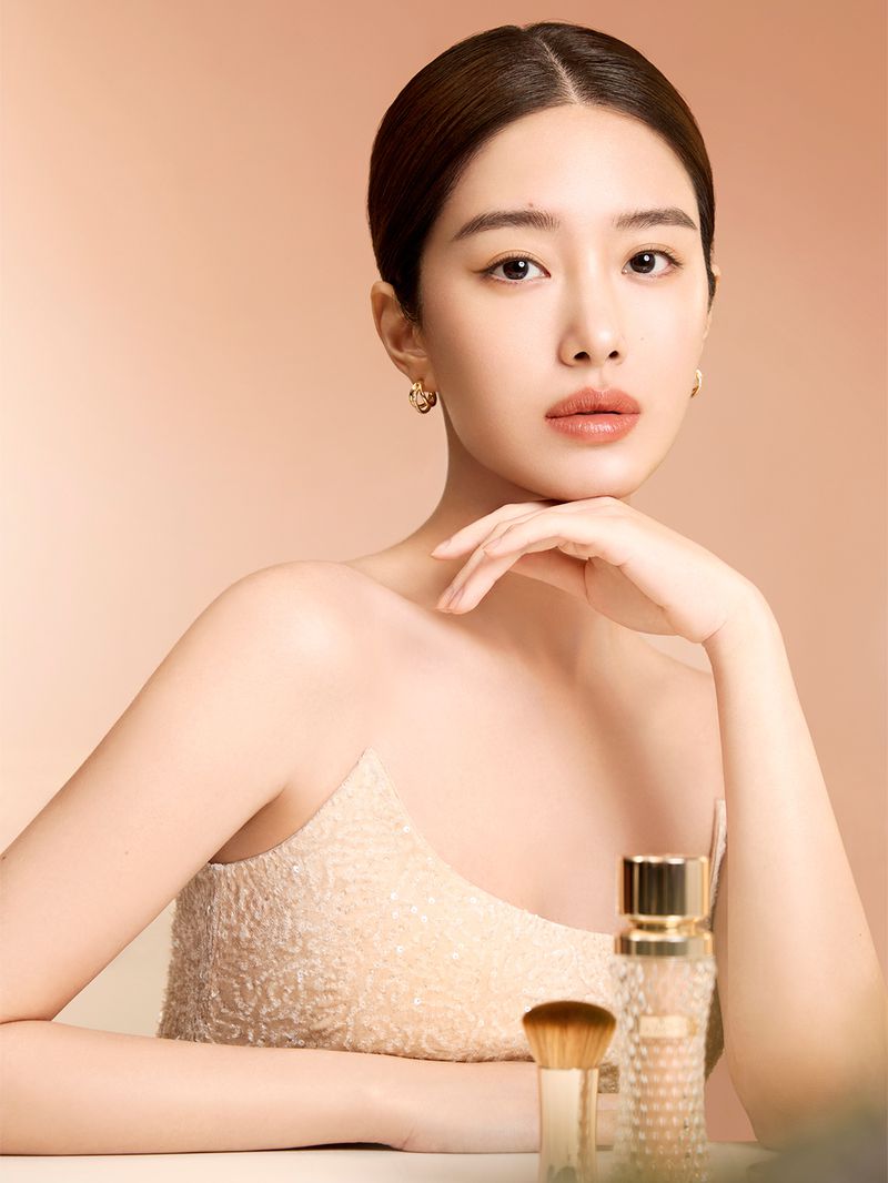 Portrait of a light-skinned adult woman with dark hair and makeup, posing with cosmetics in a studio with a peach-colored backdrop.