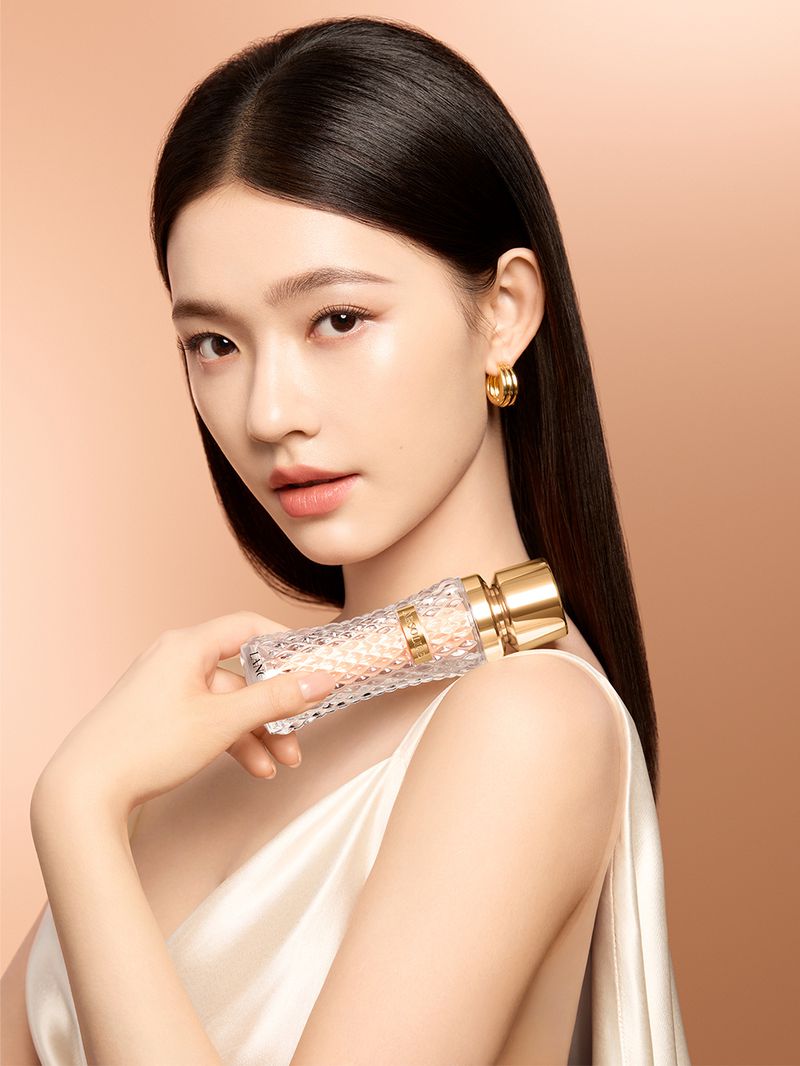 Asian woman holding faceted perfume bottle on shoulder against tan gradient background in beauty-fashion portrait. Beauty shot of perfume.