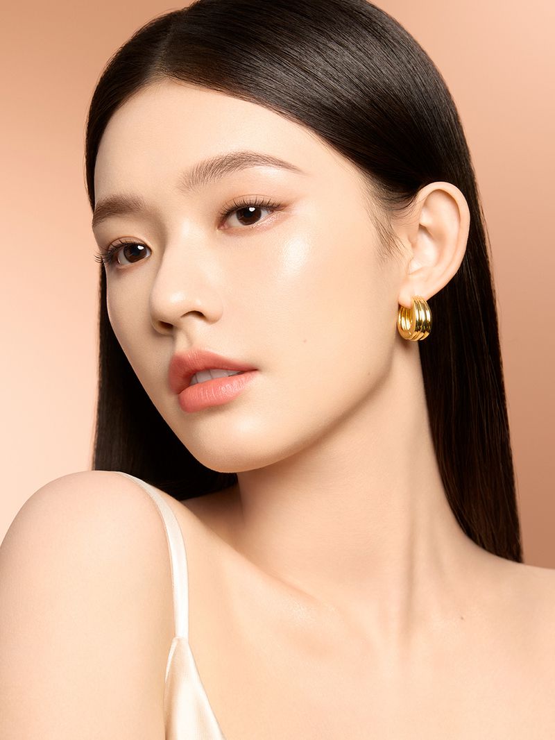 Close-up beauty shot of a woman with fair skin wearing gold hoop earrings and a cream-colored satin top against a peach background.