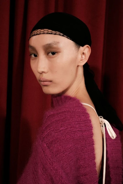 Headshot of an Asian fashion model with black headscarf posing against a red curtain backdrop, wearing a pink mohair sweater and white straps.