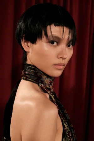 Model with short black hair and patterned neck scarf standing against a dark red curtain backdrop