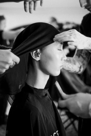 Monochrome portrait of a model getting ready backstage with multiple hands adjusting a black head covering in a fashion setting.