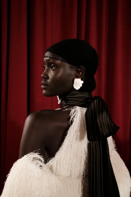 Profile portrait of a black female model in a cream, fringed dress with a black head covering against a dark-red curtain backdrop.