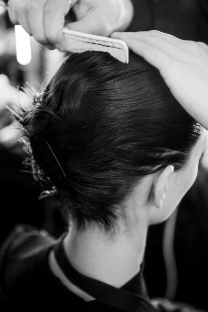 Black and white shot of a person having their dark hair styled with a comb, close-up on the back of their head and shoulders.