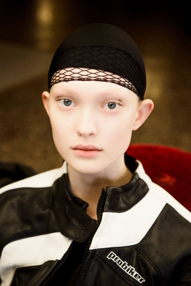 Close-up of a fair-skinned young adult female wearing a black hair net and a black and white leather jacket in an indoor setting.