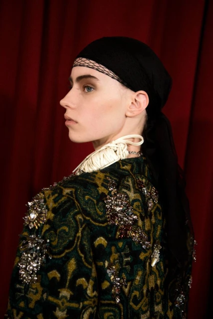 Profile of a woman wearing a black headscarf and ornamental coat with a dark red background, showcasing fashion and editorial style.