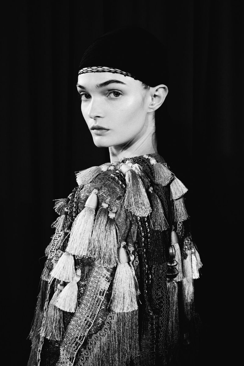 Monochrome portrait of a woman with dark headwear and an ornate tassel-detailed top, gazing toward the viewer in a dark studio.