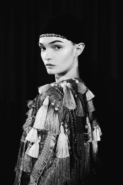 Monochrome portrait of a woman with dark headwear and an ornate tassel-detailed top, gazing toward the viewer in a dark studio.