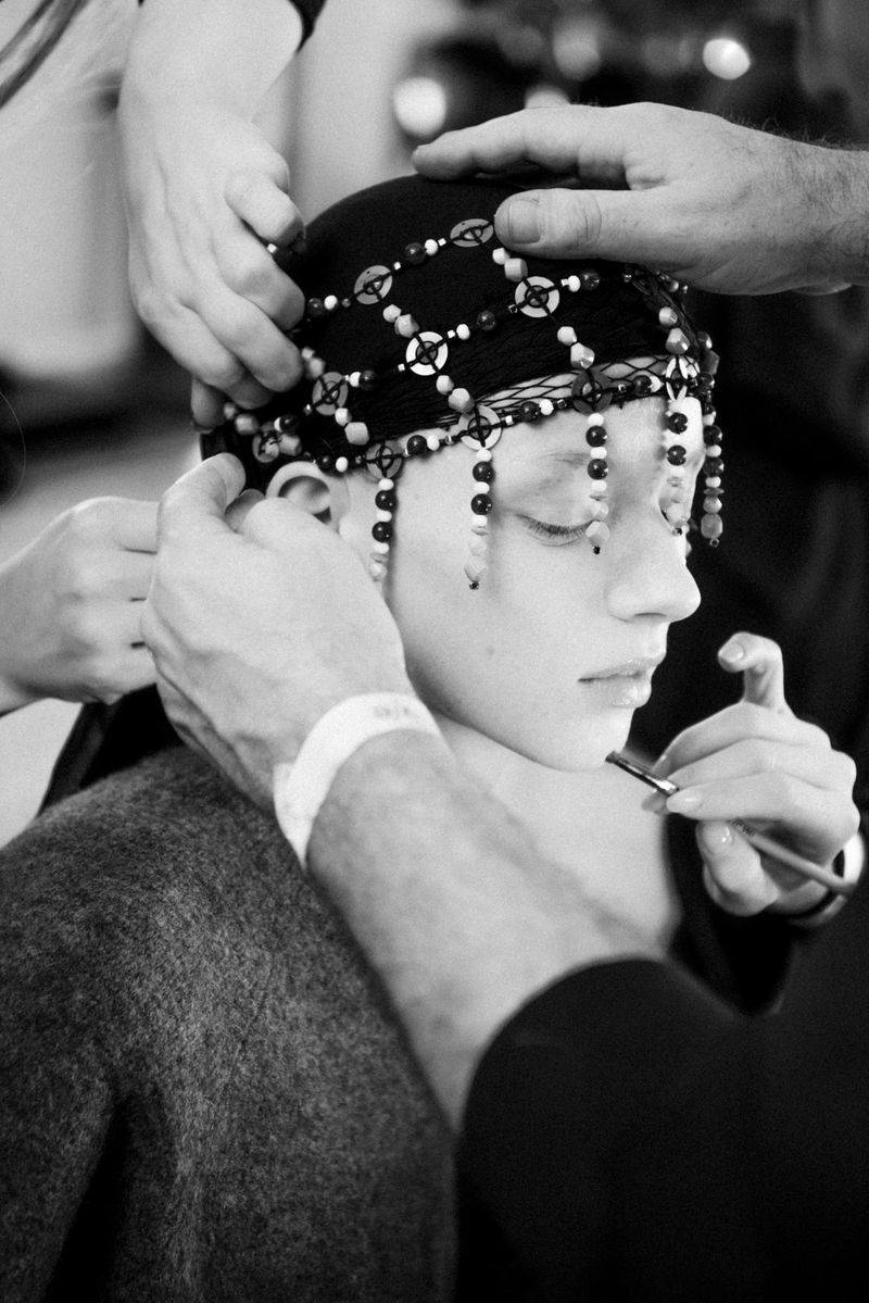 Adult female having makeup applied and beaded headpiece adjusted by multiple stylists, in a black and white backstage fashion shot.