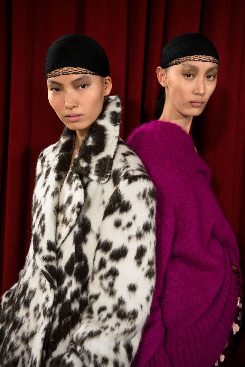 Two women in fashionable outfits pose in front of a red curtain; one wears a dalmatian-print coat, the other a purple sweater.