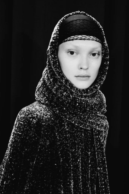 Monochrome portrait of a fair-skinned adult female model with a head covering and textured hooded cape against a dark backdrop.