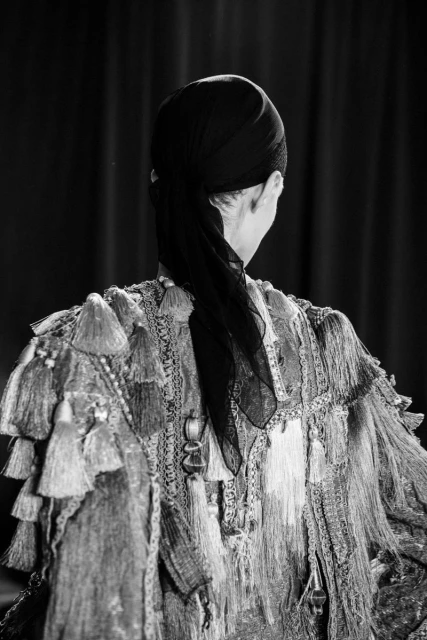 Back view of a person wearing a headscarf and an ornate garment with tassels, captured in a black and white photograph.