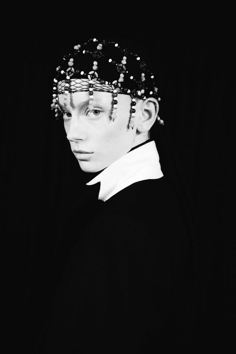 Monochrome portrait of a young adult wearing a beaded headpiece with a white collared shirt against a dark background.  
