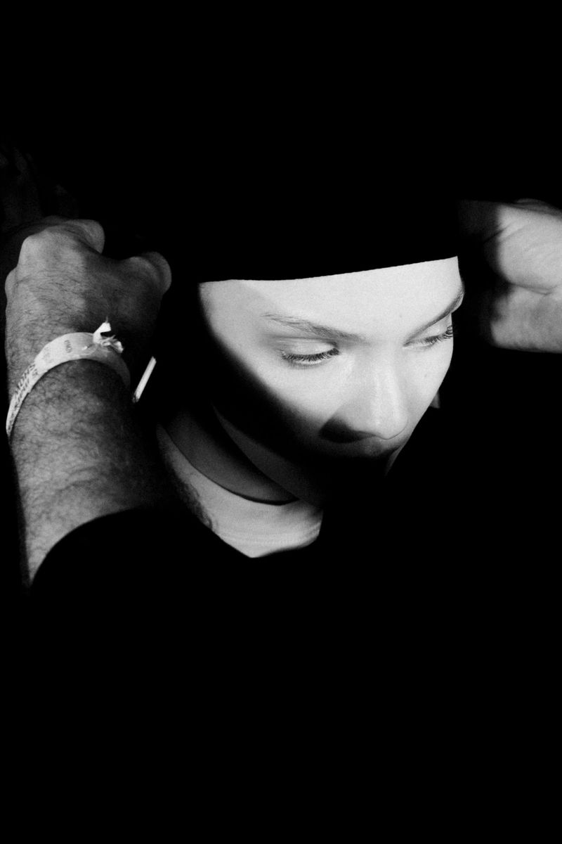 Close-up portrait of a model adjusting a dark head covering with hands visible in harsh high contrast lighting.
