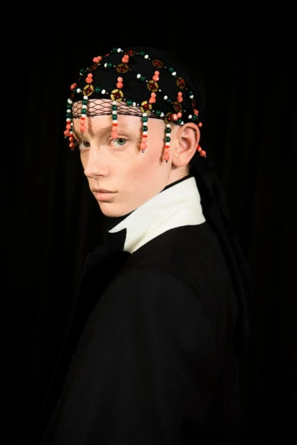 Portrait of a person with beaded headwear, white collared shirt, and dark jacket against a black background, showcasing fashion and style.