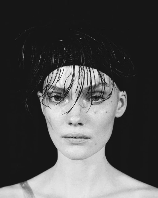 Black and white portrait of a woman with a feather headband looking directly at the camera, close-up shot against a dark background.