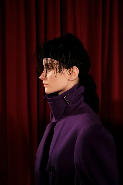 Profile portrait of a woman with dark hair and a purple coat against a dark red curtain backdrop in a fashion editorial style.