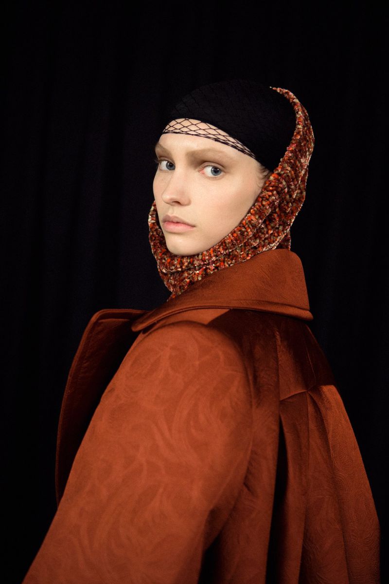 Model wearing a structured rust coat and headscarf against a dark background for Dries Van Noten F/W 25 Backstage