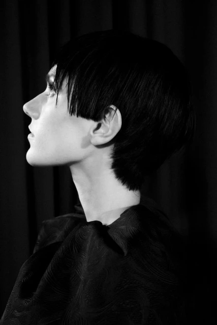 Monochrome profile portrait of a person with a short dark blunt hairstyle and wearing a dark patterned top against a dark backdrop.