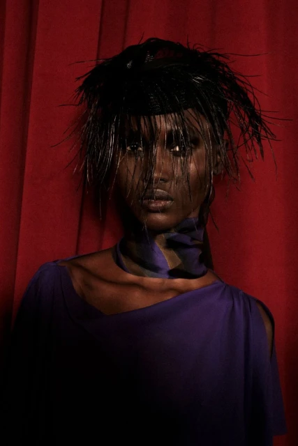 Dramatic close-up portrait of a woman with dark skin wearing a feathered hat and purple dress against a red curtain backdrop in studio lighting.