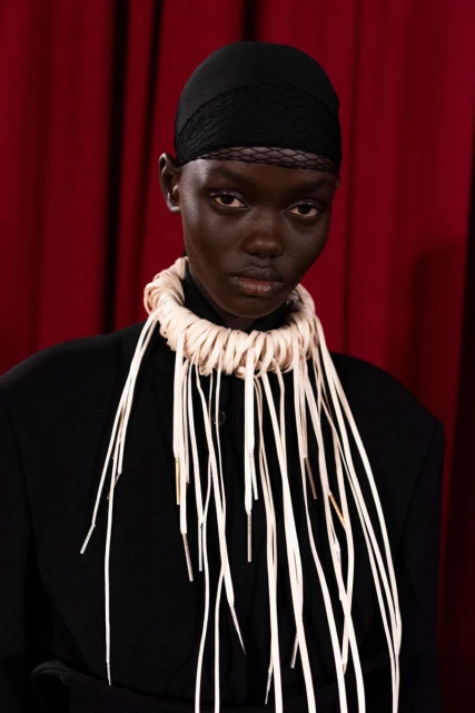 Close-up of a model in a black head covering and jacket, wearing a white decorative shoelace necklace, posing against a red backdrop.