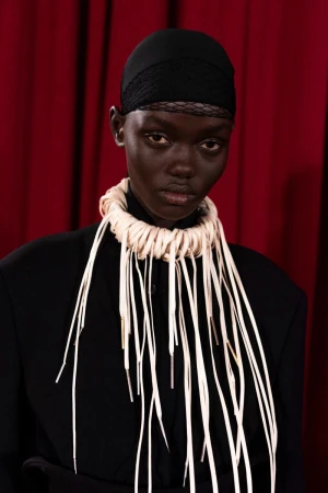 Close-up of a model in a black head covering and jacket, wearing a white decorative shoelace necklace, posing against a red backdrop.