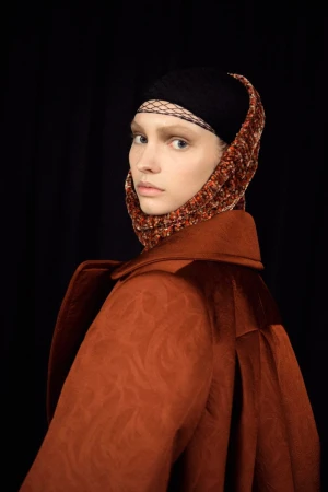 Model wearing a structured rust coat and headscarf against a dark background for Dries Van Noten F/W 25 Backstage