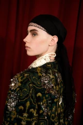 Model wearing a black headscarf and embellished green patterned top against a dark red curtain backdrop.