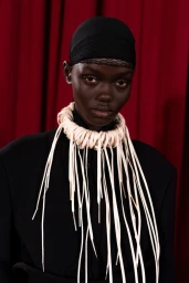 Model wearing a black cap with mesh detail and a sculptural cream-colored fringed necklace against deep red curtains.