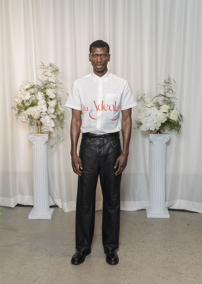 Full shot of a single adult male posing in front of a white backdrop with floral arrangements wearing black leather pants and white shirt.