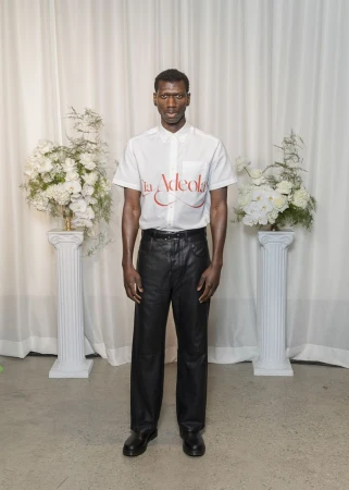 Full shot of a single adult male posing in front of a white backdrop with floral arrangements wearing black leather pants and white shirt.