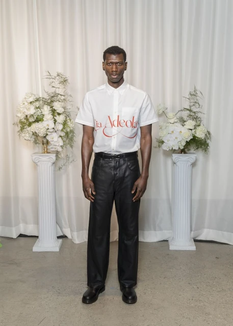 Model standing in white short-sleeve shirt and black trousers against a draped curtain background with floral pillars.