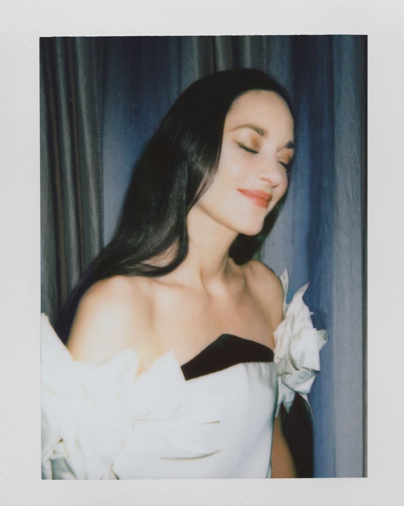 A woman with long dark hair and red lipstick is wearing a white strapless dress with a black accent and smiling with her eyes closed.