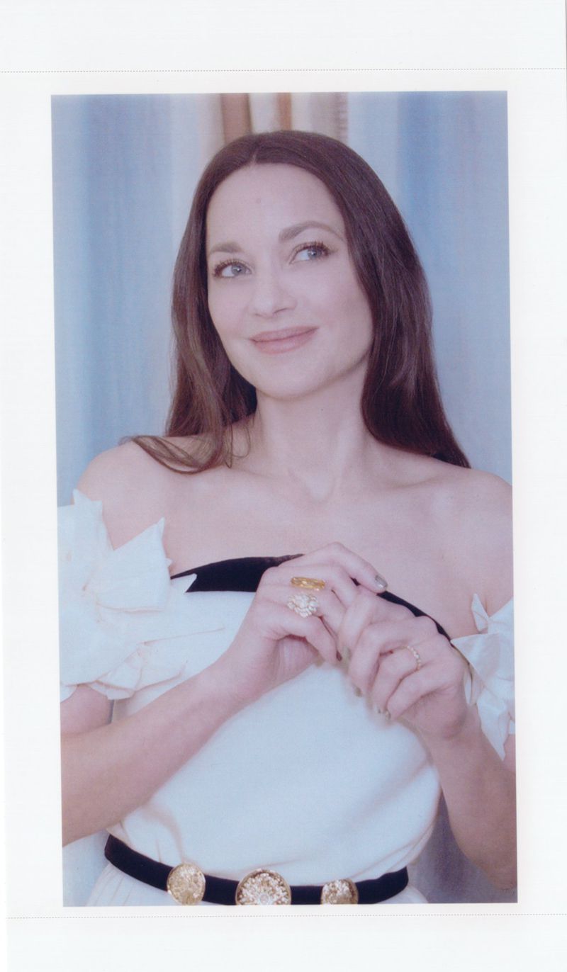 Portrait of a woman with long brown hair wearing a white off-the-shoulder dress with a black belt and ornate buckle, indoors, in soft lighting.