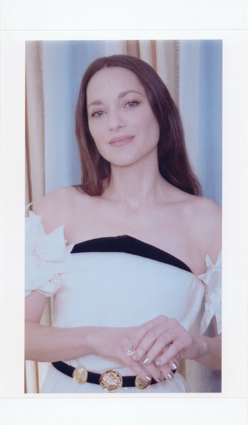 Close-up portrait of a woman with long brown hair wearing an off-the-shoulder white dress with black trim and a decorative belt, indoors.