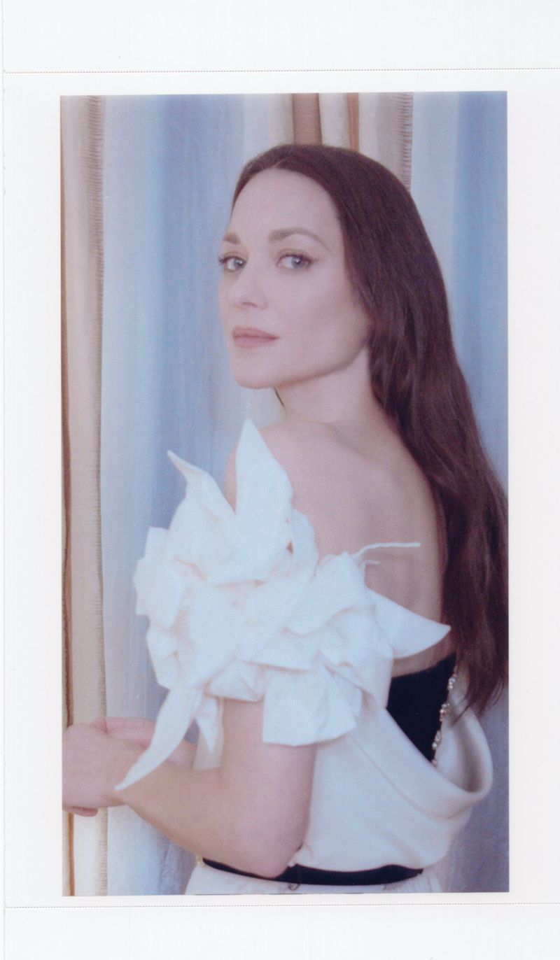 Brunette woman in a formal off-shoulder cream dress with a large ruffled sleeve, looking over her shoulder in an indoor studio setting.
