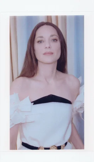 Portrait of Marion Cotillard wearing a white off-the-shoulder dress with black trim against vertical striped curtains