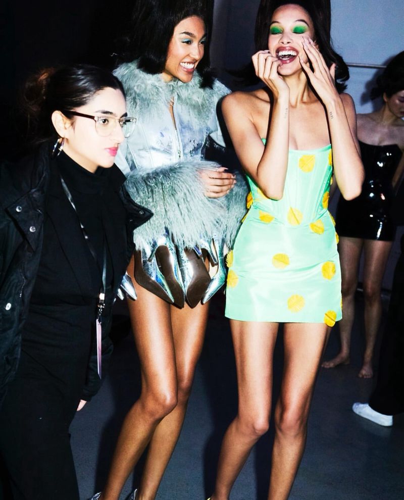 Three women pose together. One wears a pale-green polka dot dress; another has silver accents and fur. A third wears a black jacket.