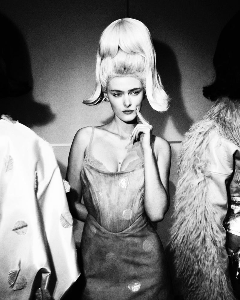 Black-and-white shot of a stylish adult female with elaborate blonde hair and unique dress, flanked by two obscured figures.