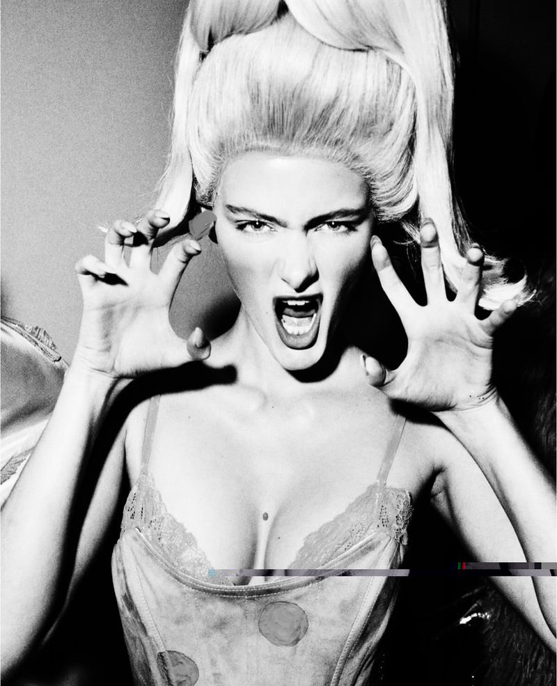 Dramatic black and white portrait of an adult female screaming with hands up, wearing a vintage corset and tall blonde wig.