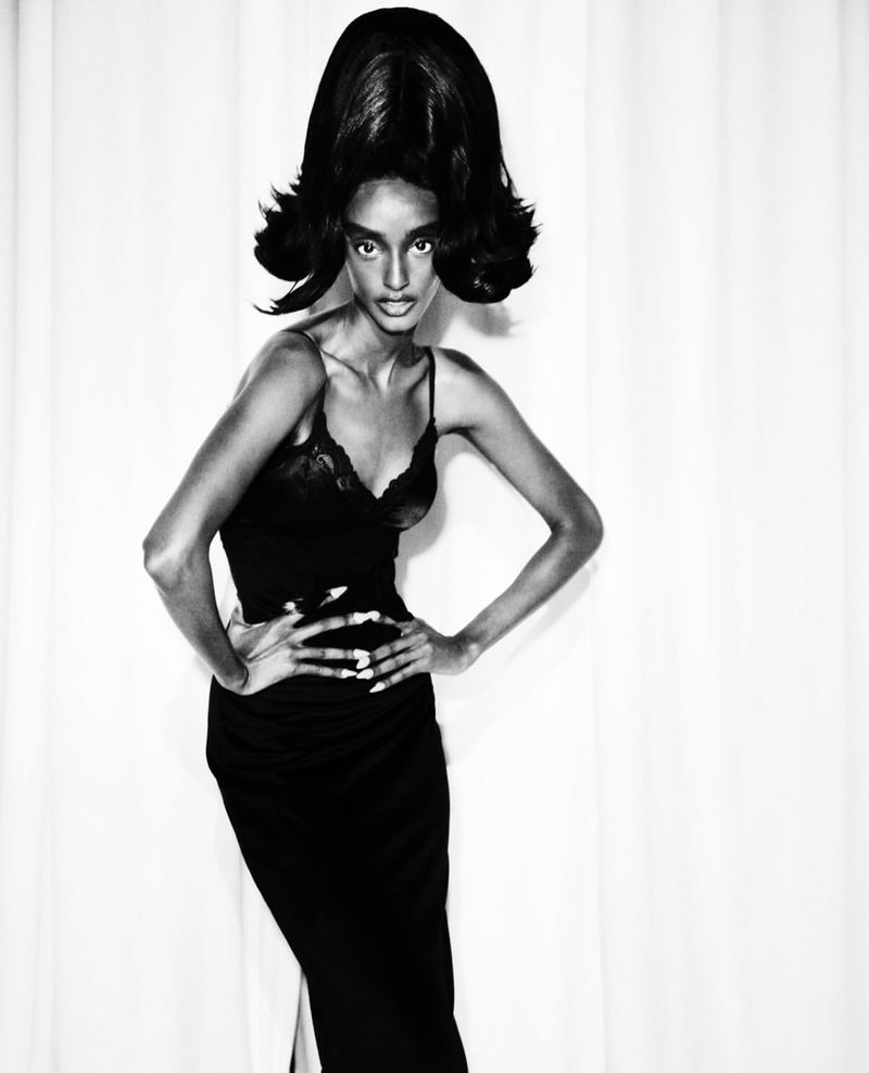 Monochrome fashion portrait of a Black woman with a voluminous beehive hairstyle, wearing a sleek black dress, against a white backdrop.