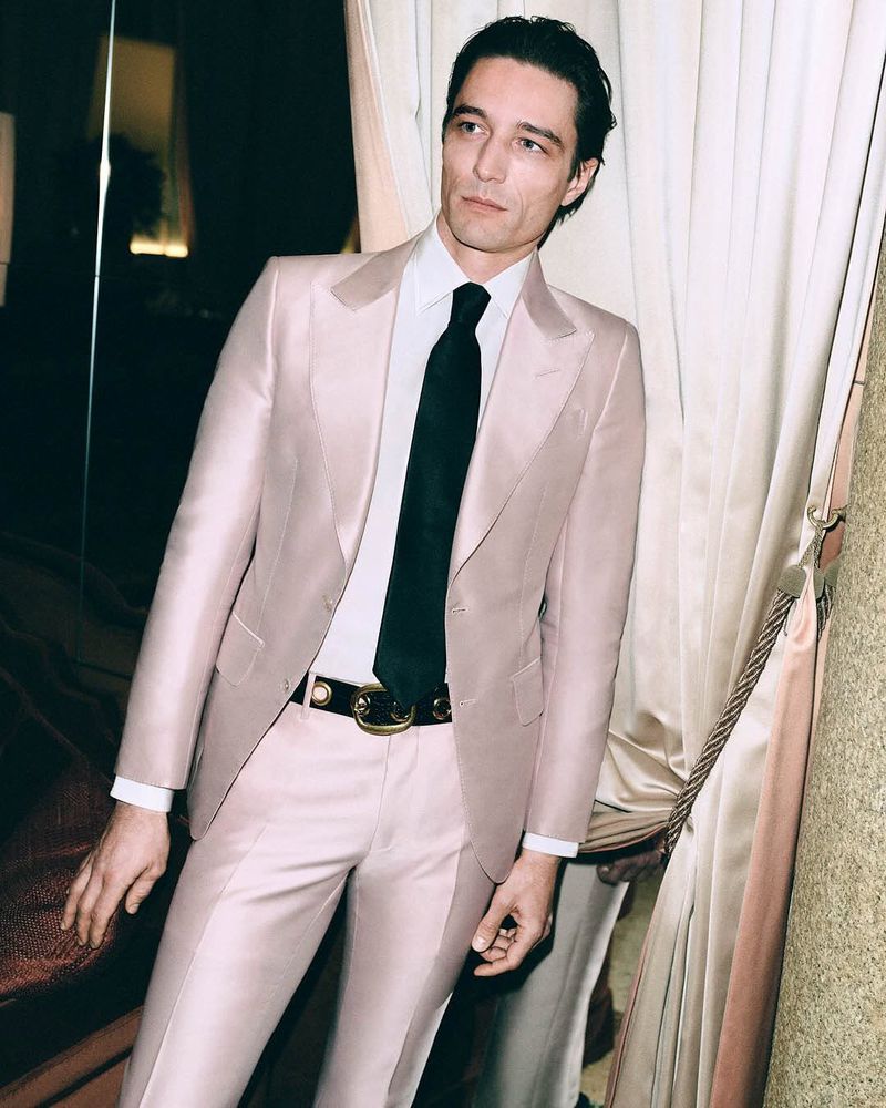 A man in a pale pink suit and black tie poses against a curtained wall in a stylish indoor portrait, showcasing fashion and elegance.