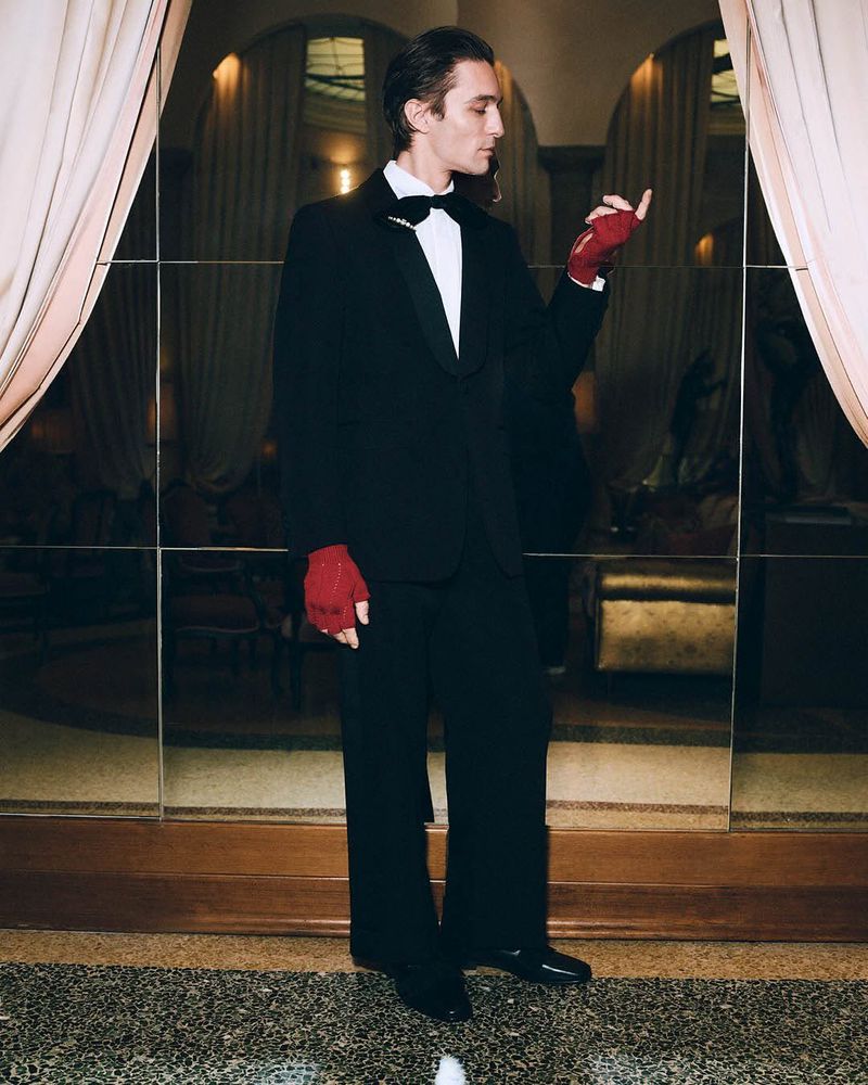 Elegant man in a black tuxedo with red gloves stands in front of mirrors and a light-colored curtain for a sophisticated fashion look.