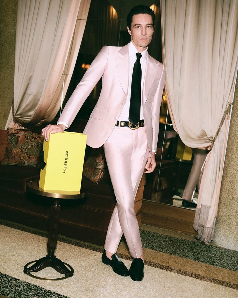 Man in a light pink suit poses indoors with a yellow gift box in a stylish setting, showcasing fashion and elegance.