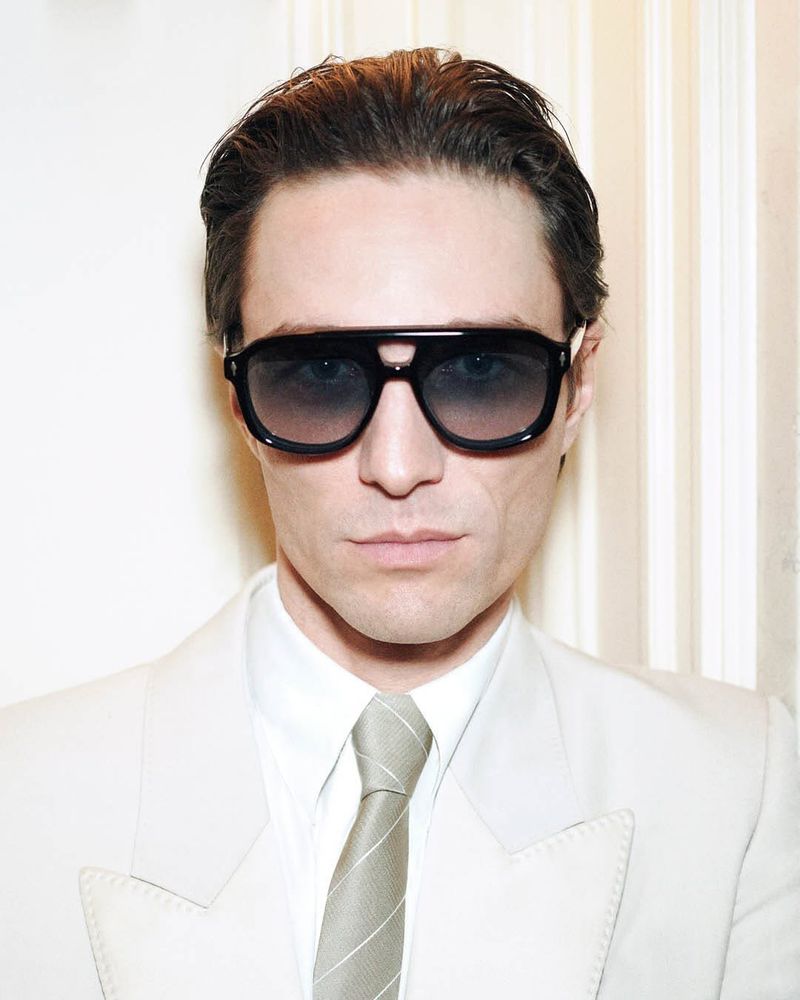 Portrait of adult male wearing sunglasses, tie, and suit jacket in front of curtained wall, shot in studio with neutral lighting.