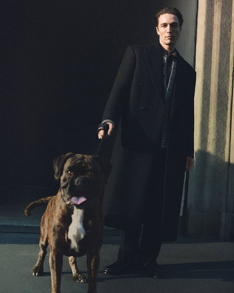 An adult male in a black coat walks a brindle boxer dog on a leash in a city with dark light against a neutral wall.