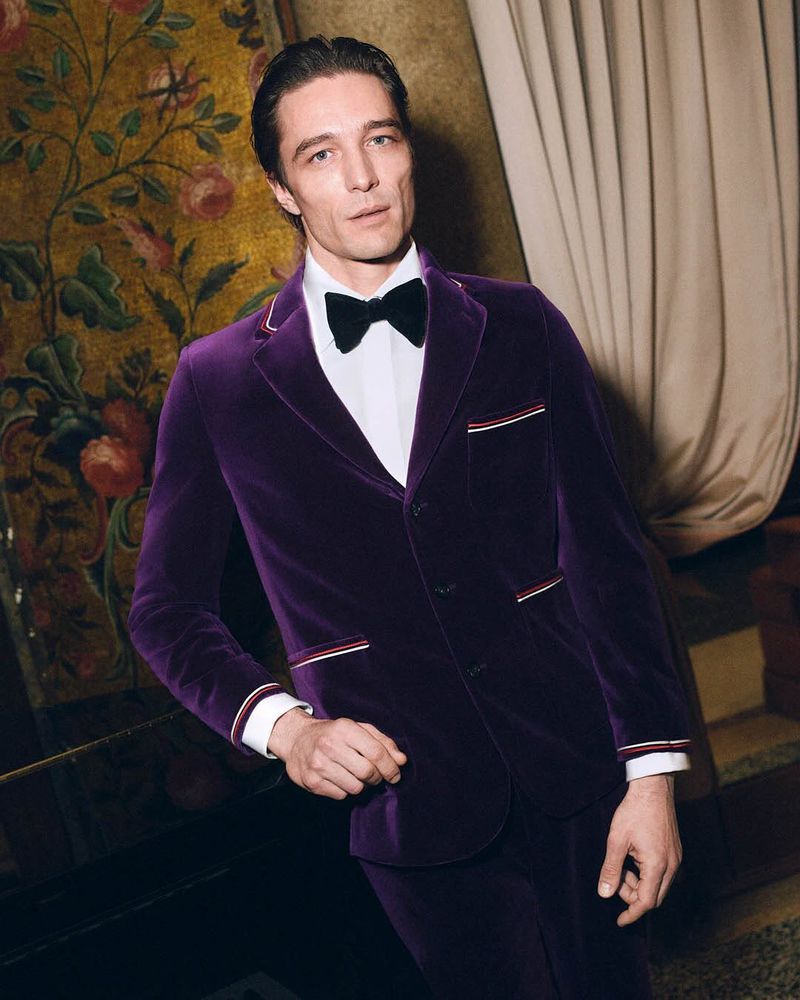 A man in a purple velvet suit and bowtie stands indoors, posing against a backdrop of patterned wallpaper in a medium shot.